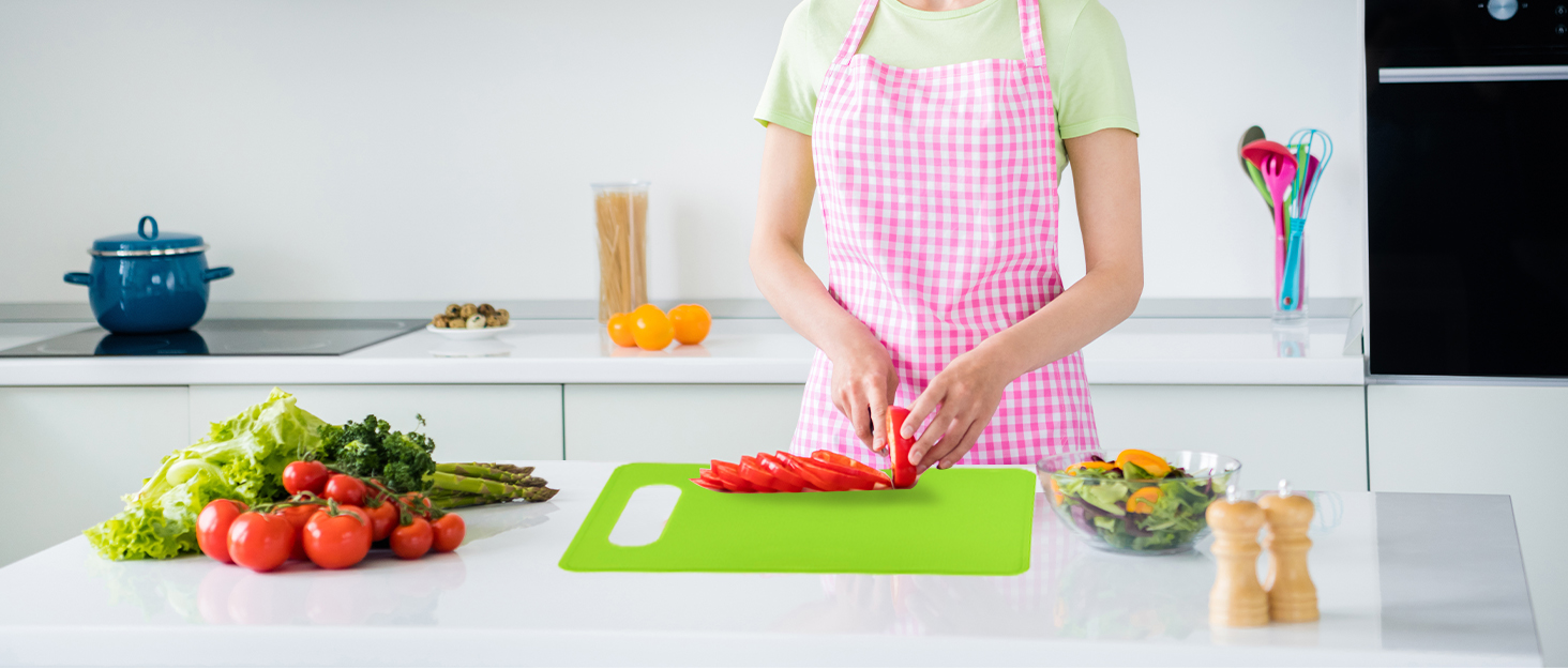 Young housewife in apron standing at the kitchen table and cutting vegetables for salad