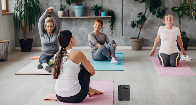 Indoor yoga or exercise session showing group on yoga mats in spacious room with plants. Smart speaker visible on floor.