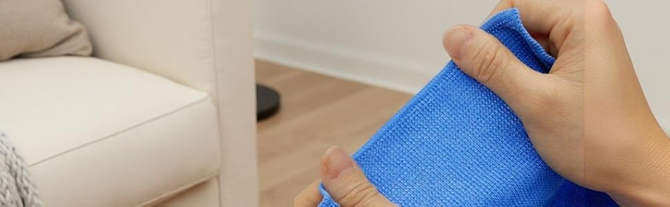 Hands folding blue microfiber cleaning cloth near beige armchair and white wall, with pink candle visible in scene.