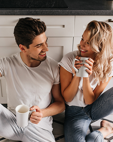 Two people relaxing in a casual kitchen setting, sharing beverages in white mugs while seated comfortably together.