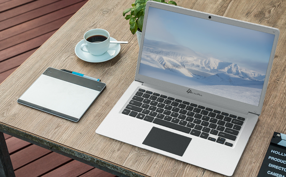 Laptop with snowy mountain wallpaper on wooden table outdoors. Coffee cup, notepad, and pen nearby. Device appears slim with metallic finish.