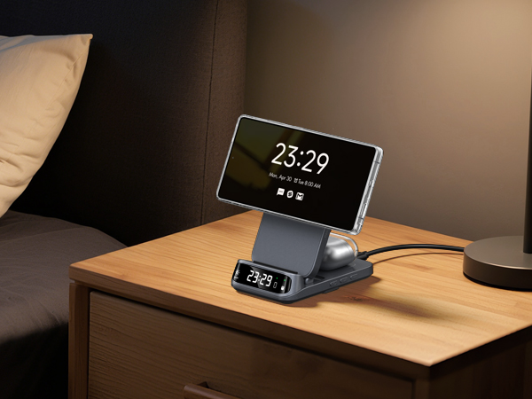 Smartphone charging dock on wooden nightstand. Device displays time 23:29 on large screen. Smaller digital clock on base. Sleek design with adjustable viewing angle.