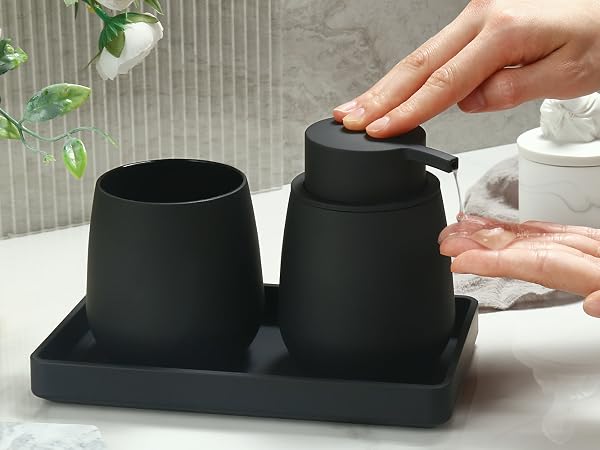 Matte black bathroom set on tray: soap dispenser with hand pumping, tumbler, and rectangular base. Minimalist design in use on counter with plant visible.