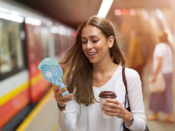 Woman holding a small handheld fan and disposable coffee cup while standing near a subway train. She appears to be cooling herself while commuting.