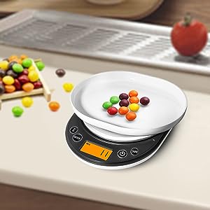Digital kitchen scale with white circular platform, displaying weight of colorful candy pieces. Stainless steel grater and red tomato visible in background.