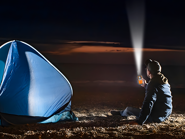 A camper sits by tent at night using walkie talkies with its flashlight feature to light darkness