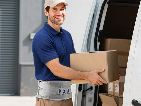 Delivery person wearing a blue polo shirt and white cap, carrying a cardboard box. They're wearing a gray back support belt with blue accents.