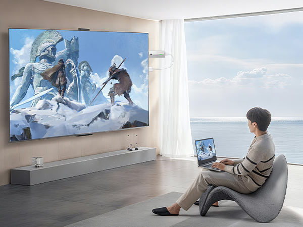 Modern living room setting with large wall-mounted display showing winter mountain scene. Person seated in curved modern chair using laptop, with ocean view through windows.